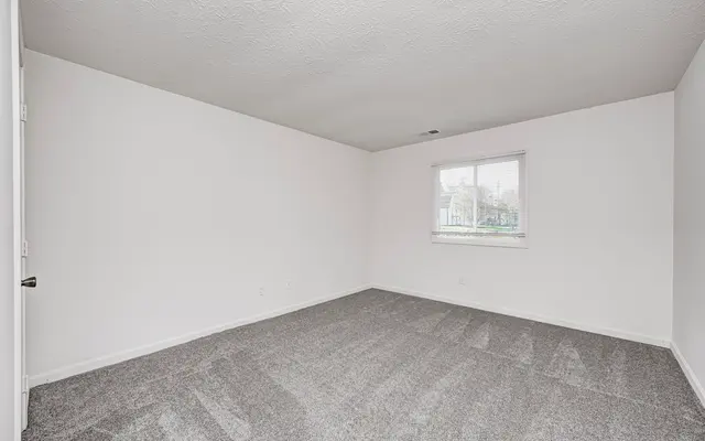 A vacant room with light gray carpet, plain white walls, and a single window letting in natural light.