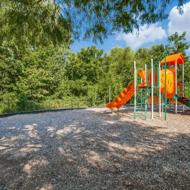 A playground featuring a colorful slide surrounded by trees and gravel flooring.