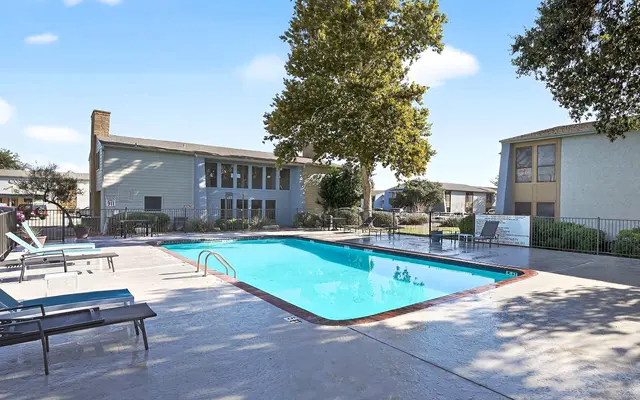 A pool area with a clear blue pool, surrounded by lounge chairs and two buildings in the background under a blue sky.
