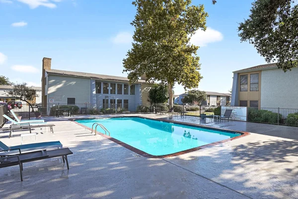 A pool area with a clear blue pool, surrounded by lounge chairs and two buildings in the background under a blue sky.
