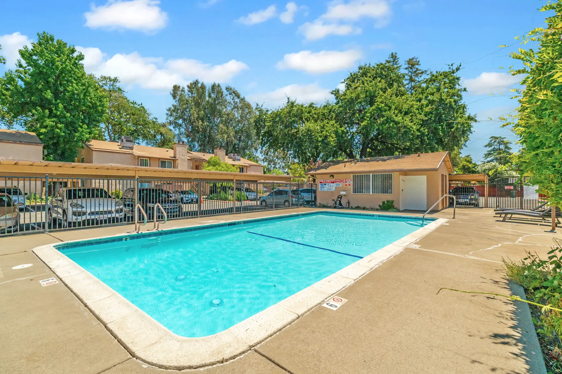 A clear blue swimming pool surrounded by greenery and residential buildings. The pool area features a safety fence, lounge chairs, and a small building nearby.