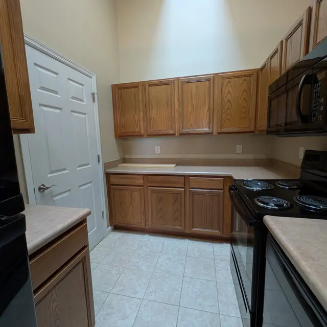 A modern kitchen featuring wooden cabinets, a black stove, and dark appliances. The walls are painted in a warm beige tone. The floor is tiled with light-colored tiles.