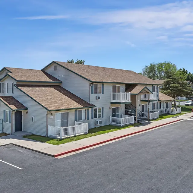 Apartment Complex Overview A view of a two-story apartment complex with light-colored exterior and brown roofs, surrounded by green grass and trees, with a parking lot in the foreground.