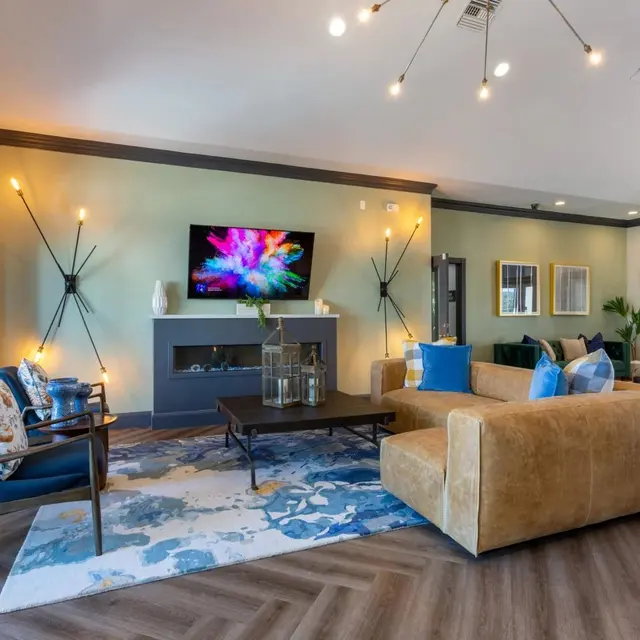 A stylish and modern living room featuring a large window, a flat-screen TV mounted on the wall, a coffee table, and comfortable seating with decorative cushions. The room has a textured area rug and stylish lighting fixtures.