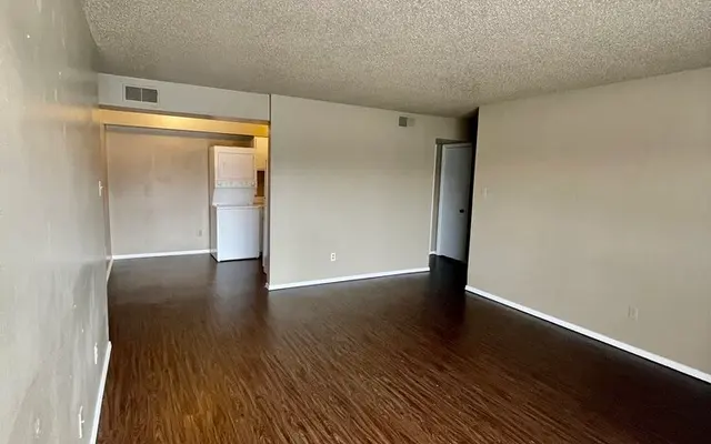 Spacious Empty Living Room A spacious empty living room with wooden flooring and light-colored walls. The ceiling is textured and sloped. There are doorways leading to other rooms, with a small kitchen visible at the far end.