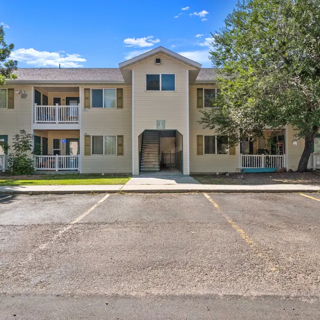 Front view of a two-story apartment building with balconies on both levels, surrounded by mature trees and landscaping. The building features a central stairway entry and shaded areas, providing a welcoming and comfortable residential environment.