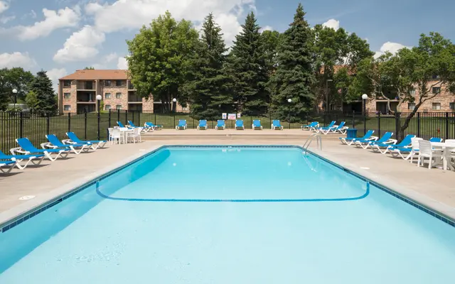 an exterior view of The Woods of Burnsville fenced swimming pool surrounded by lounge chairs and a view of the apartment buildings and trees - Hotel, Building, Pool