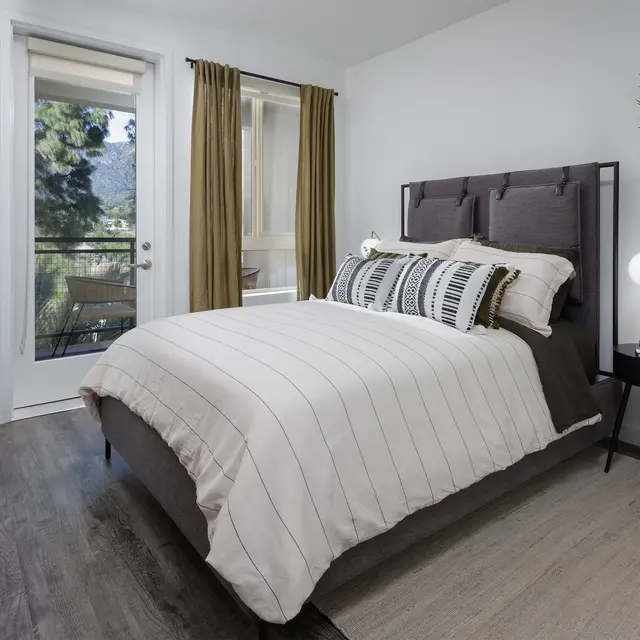 A modern bedroom featuring a bed with striped bedding, a large window with curtains, and a potted plant in the corner.