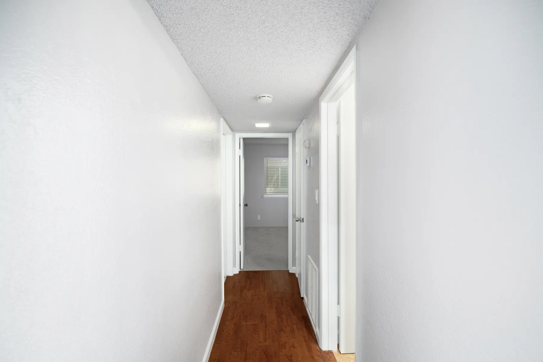 A long, brightly lit hallway with white walls and a wooden floor. Two doors are visible on the right side, leading to separate rooms, and a window at the end of the hallway allows natural light to enter.