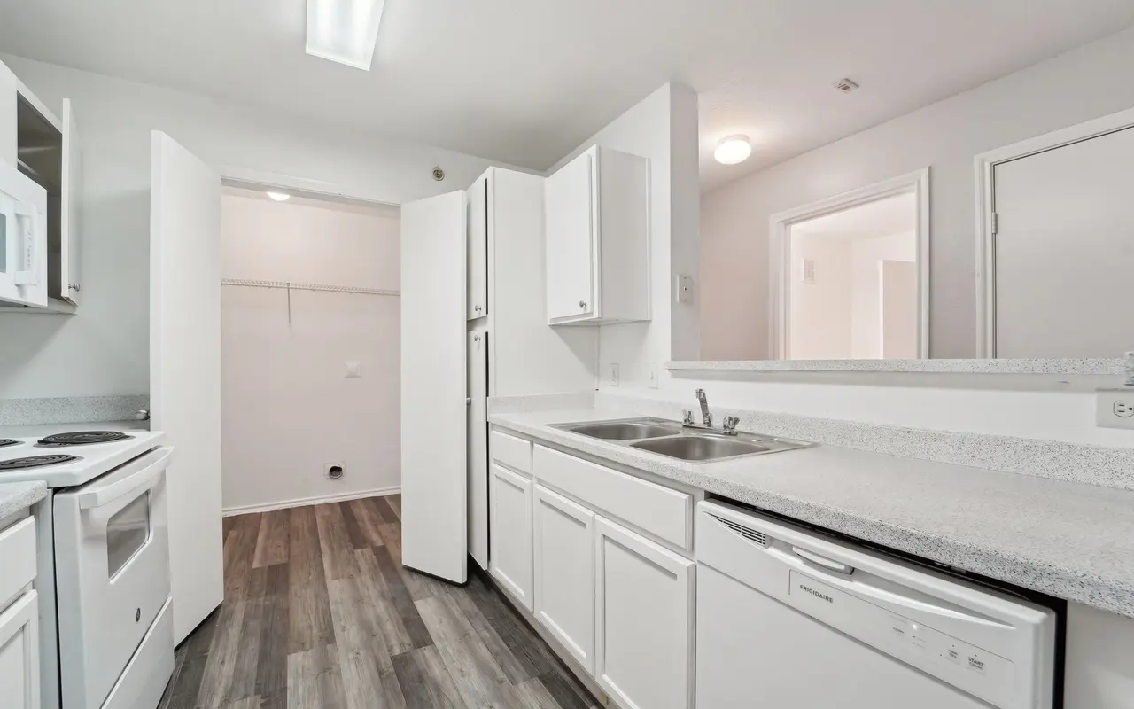 A modern kitchen with white cabinetry, gray countertops, and appliances including a stove, dishwasher, and microwave. The floor is covered with plank-style flooring and there is a doorway to a room on the right.