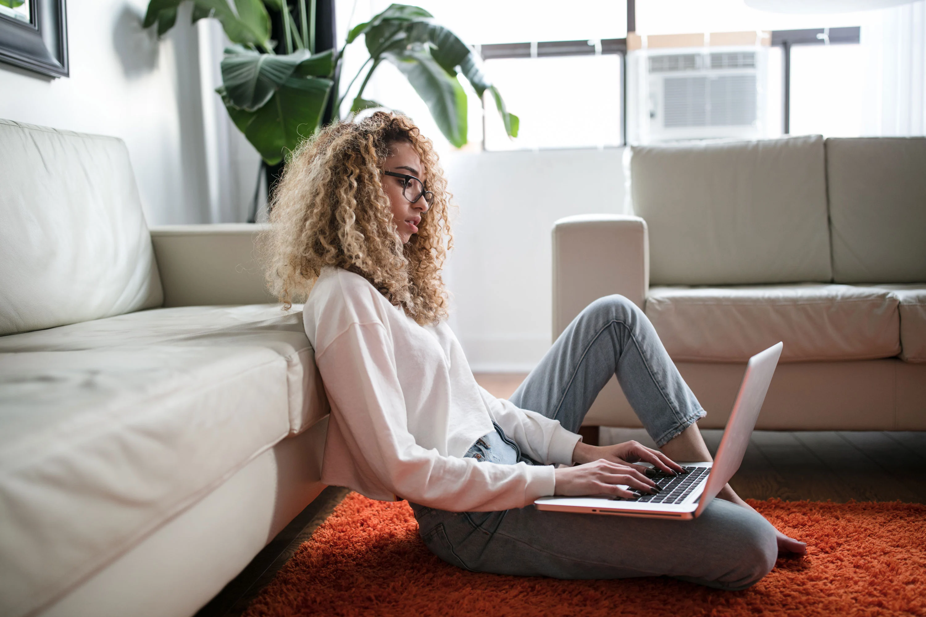 A person with curly hair sits on a carpet, typing on a laptop while leaning against a sofa in a bright room.