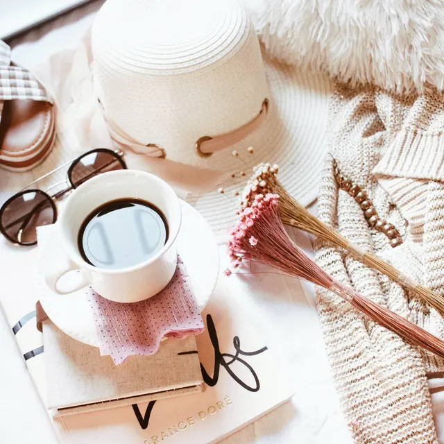 A cozy flatlay with a coffee cup, sunglasses, hat, sweater, flowers, and a book on a light fabric background.