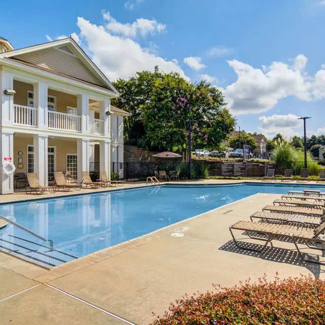 A swimming pool area featuring a large pool with sun loungers surrounding it, against a backdrop of residential buildings and trees under a partly cloudy sky.
