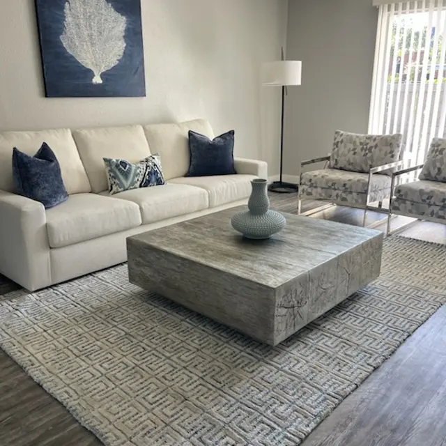 A contemporary living room featuring a white sofa with blue decorative pillows, a large square coffee table made of wood, patterned area rug, and two patterned armchairs. A piece of art with a shell design hangs on the wall, and there is a floor lamp in the corner.