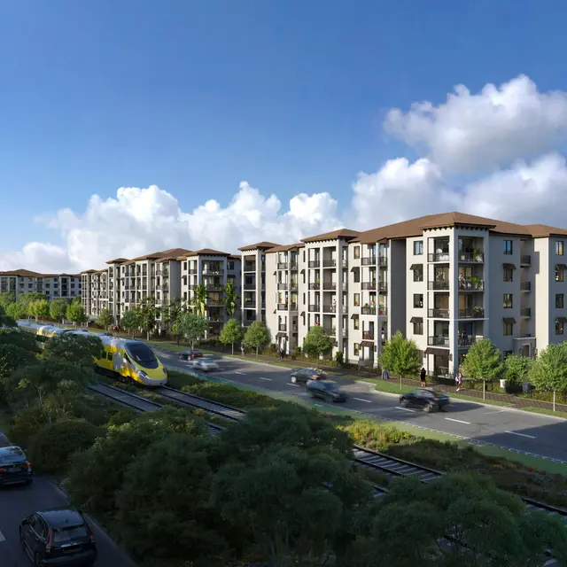 A modern apartment complex features multi-story buildings with balconies, situated next to a roadway and railway tracks, surrounded by greenery under a blue sky with clouds.
