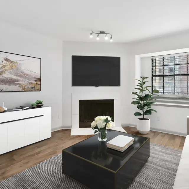 A modern living room featuring a white wall, a mounted flat-screen TV, a sleek black coffee table, and a white sideboard. There is a green plant in a pot near a window and an artwork on the wall.