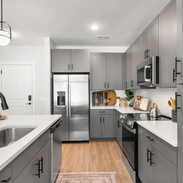A modern kitchen featuring gray cabinets, stainless steel appliances, and a white countertop with a large sink. The space includes a dining area with decorative items and a warm wood floor.