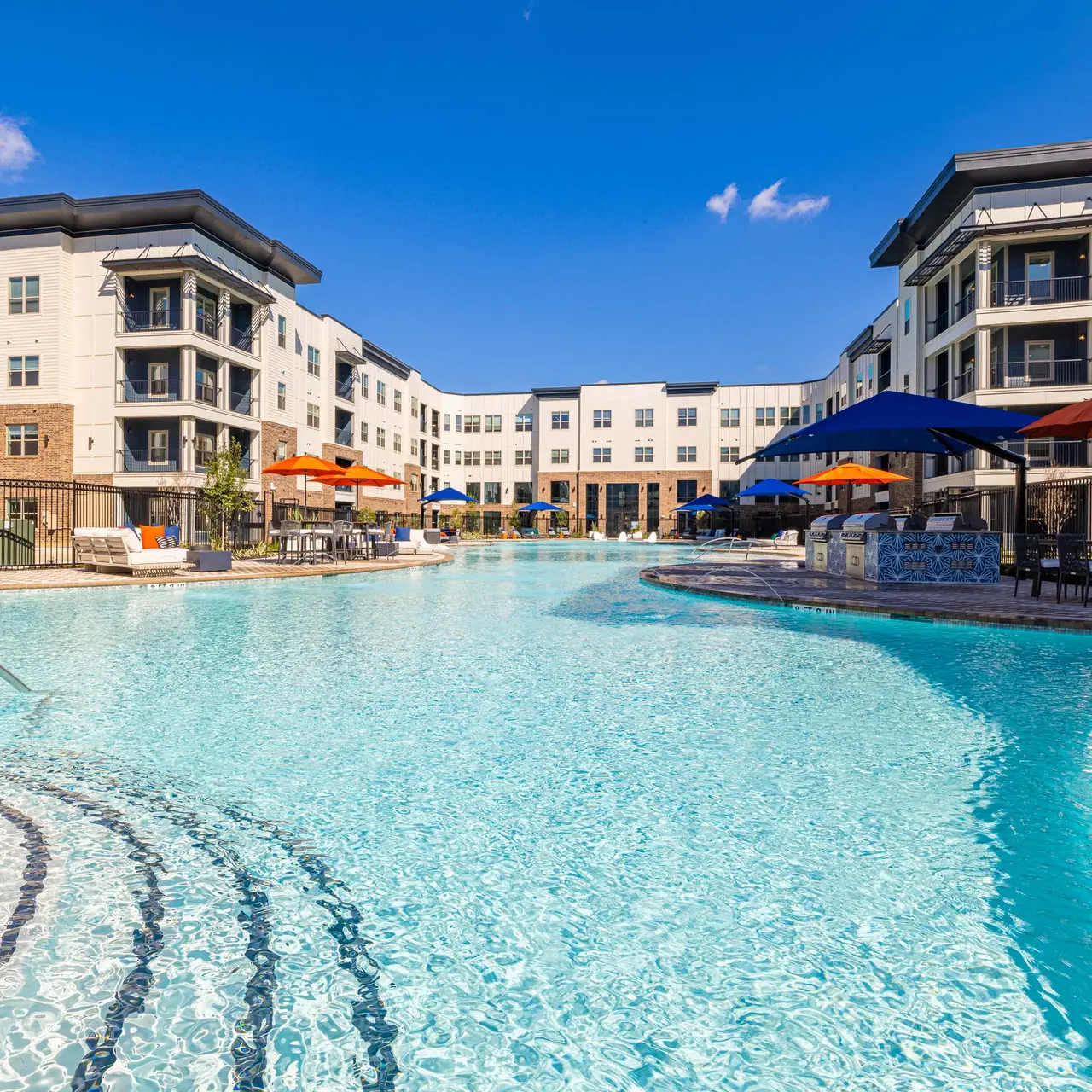 A spacious swimming pool surrounded by modern residential buildings. The pool features lounge chairs, umbrellas, and a clear blue sky overhead.