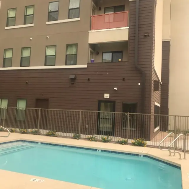 Modern Apartment Pool Area A view of a swimming pool in a residential area with an apartment building in the background. The building features multiple windows and balconies, some with railings. The pool area is surrounded by a fence, with a few potted plants along the poolside.
