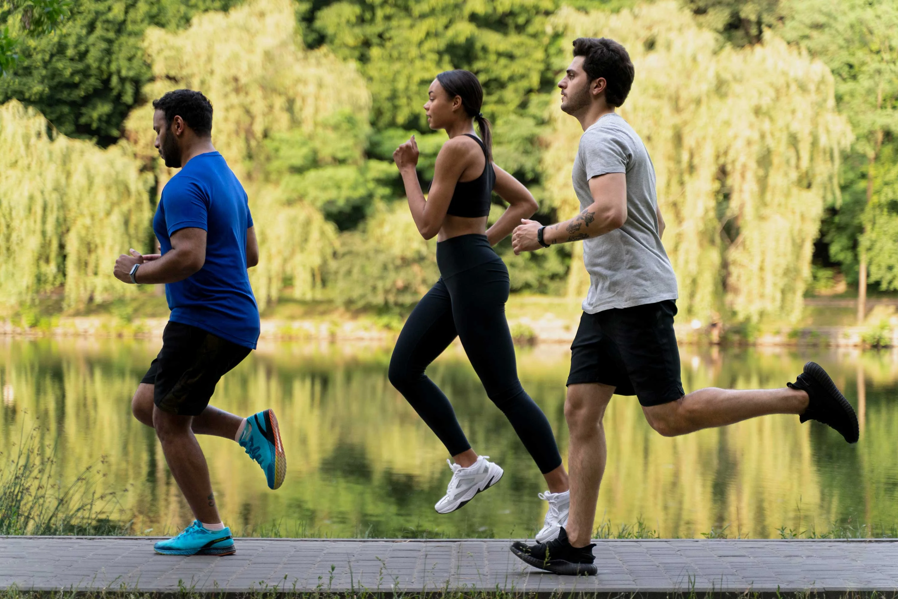 Three individuals running along a path near a body of water surrounded by trees.