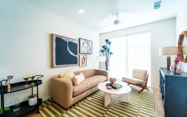 Contemporary Living Room Design A modern living room featuring a beige sofa, white coffee table, and a wooden side chair. The room is adorned with artwork on the wall and a decorative plant near the window. A stylish bar cart is placed in one corner.