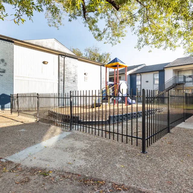 A fenced playground area with equipment, surrounded by two residential buildings.