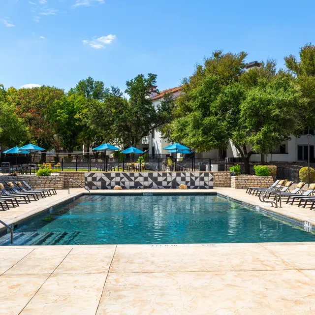 A serene pool area with a clear blue swimming pool surrounded by comfortable lounge chairs. The area is enclosed by lush green trees and blue umbrellas, with residential buildings in the background.