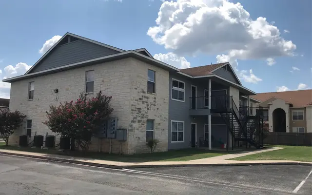 Exterior view of a two-story apartment building with a mix of brick and siding, surrounded by grass and trees under a partly cloudy sky.