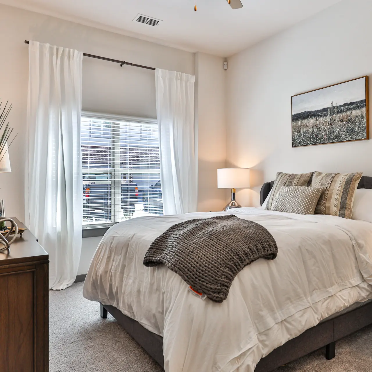 Cozy Modern Bedroom Interior A modern bedroom featuring a bed with white bedding, a textured throw, two bedside tables with lamps, and a large window with white curtains. A piece of art hangs above the bed, and there is a dresser on the left with decorative items and a potted plant.