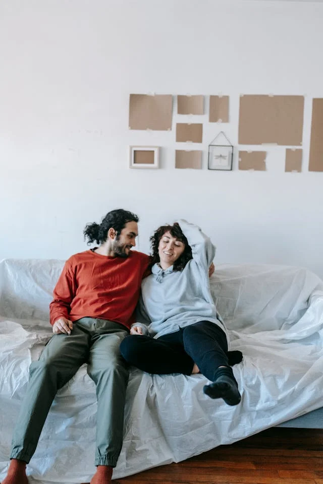 Cozy Couple Moment on a Sofa A couple sitting closely together on a couch, smiling and enjoying each other's company. The male is wearing a red sweater and green pants, while the female is in a gray hoodie.