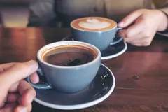Two cups of coffee on a wooden table, one being held by a hand.