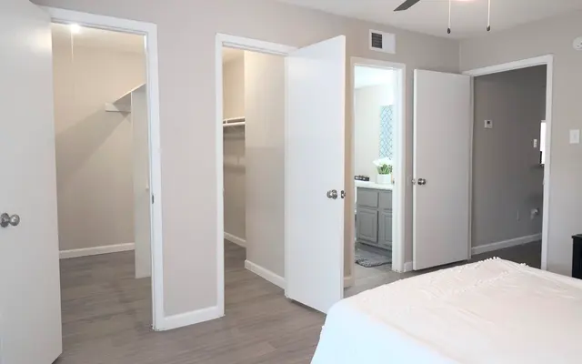 A spacious modern bedroom featuring several open white doors leading to different areas. The room has light gray walls and wooden flooring with a bed partly visible in the foreground.