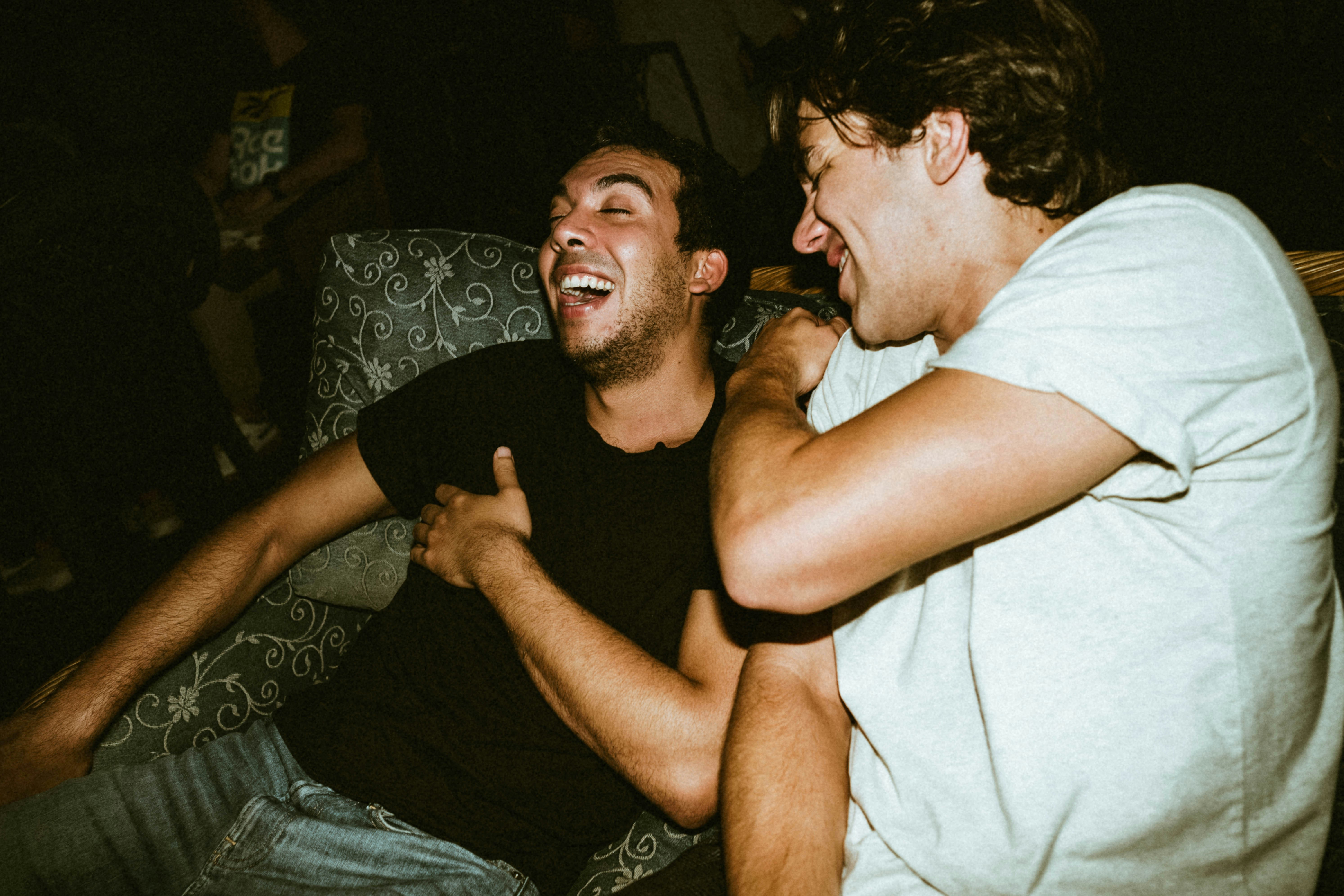 Two young men laughing together while sitting on a couch in a dimly lit setting.