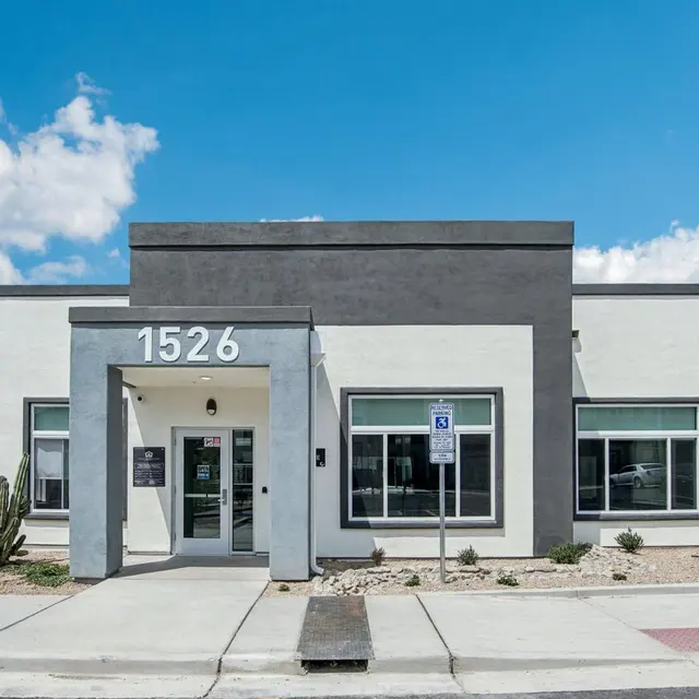 A modern, single-story commercial building with a gray and white exterior, featuring large windows and a front entrance. The building has landscaping with small cacti and is set against a blue sky with clouds.