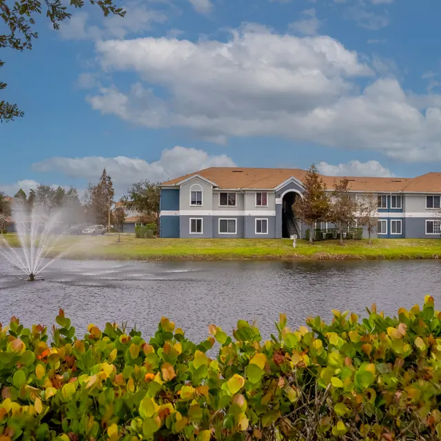 A scenic view of an apartment complex near a pond with a fountain. In the foreground are green bushes, and the background features a blue sky with scattered clouds.