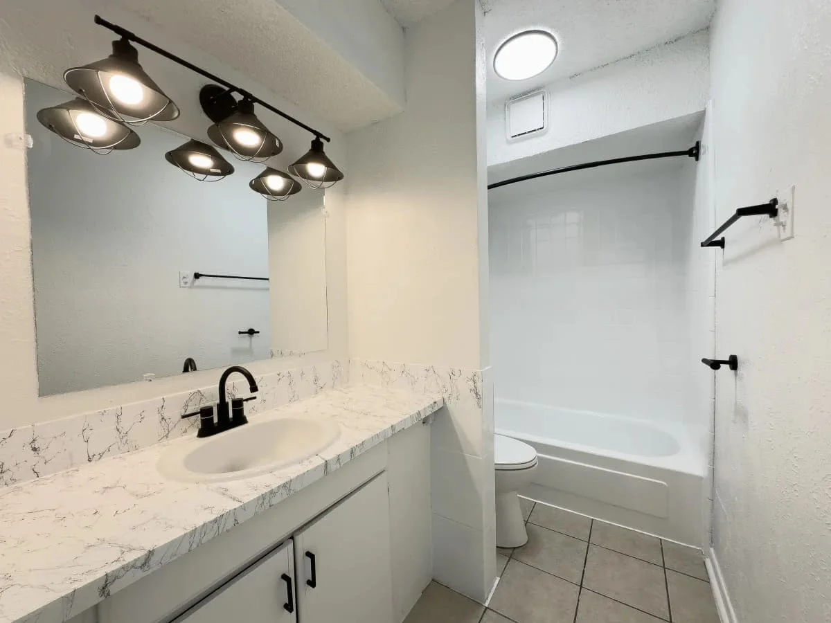 Modern Bathroom Design A modern bathroom featuring a marble countertop with a white sink, a large mirror, and stylish lighting fixtures. There is a white bathtub with a shower curtain, and neutral-colored tiles on the floor.