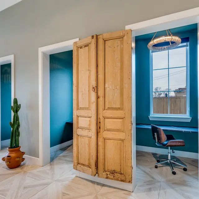A modern office interior featuring multiple doorways with wooden shutters, a cactus in a pot, and a desk area behind one of the doors. The walls are painted in a soothing blue and light gray color scheme, with elegant tiled flooring.