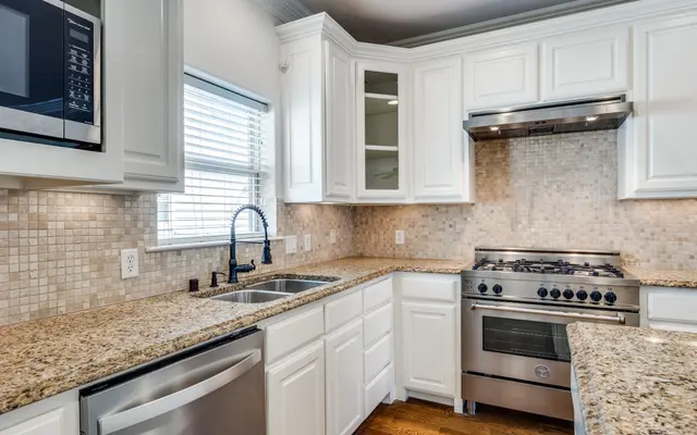 Modern Kitchen Interior A modern kitchen featuring white cabinetry, a granite countertop, a stainless steel gas stove, and a sleek faucet. The backsplash consists of light-colored tiles, and there is a window allowing natural light to enter.