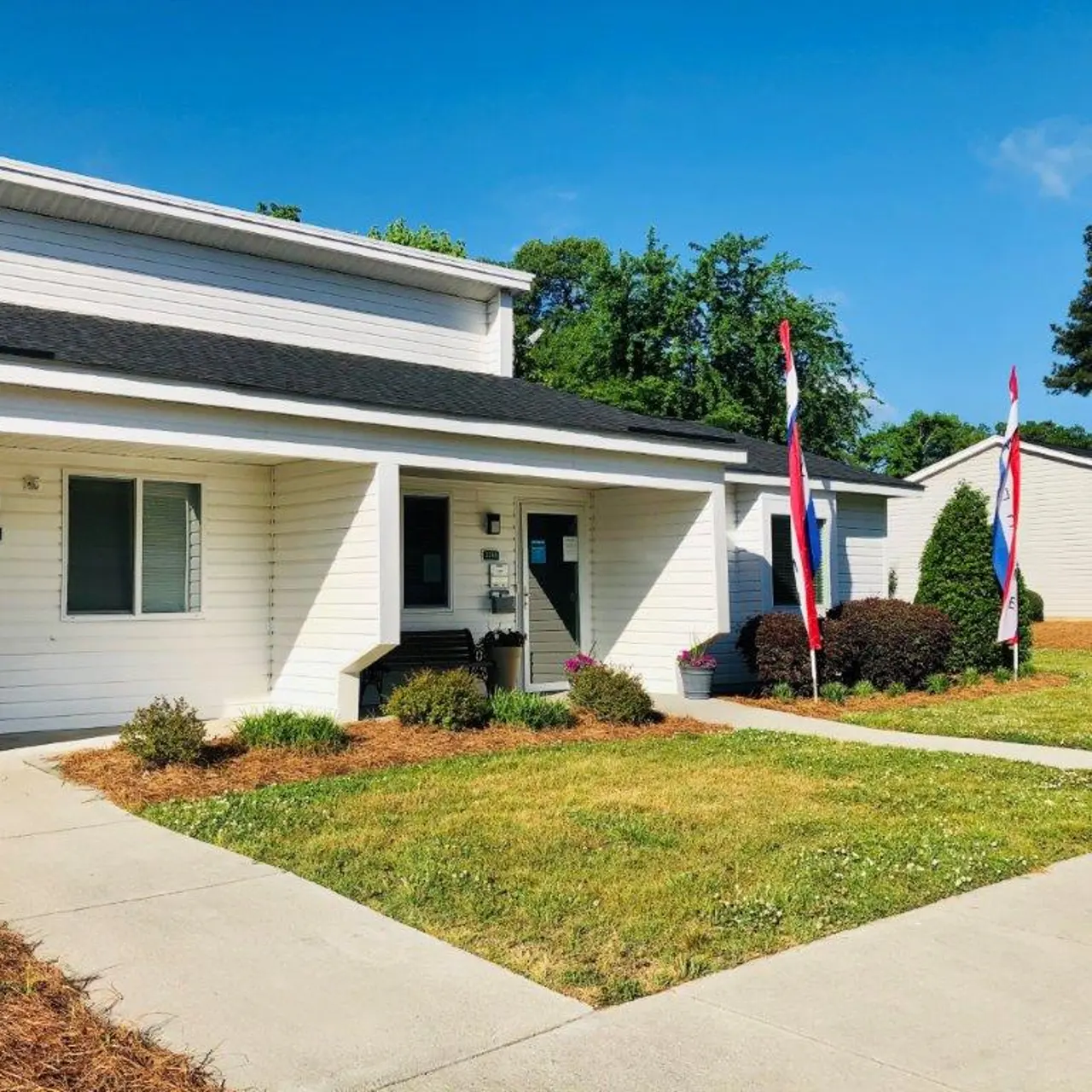 A row of single-story modern buildings in a residential neighborhood, featuring green lawns and decorative flags.