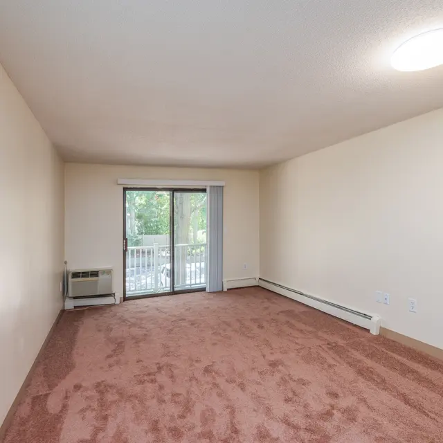 A vacant living room with light beige walls and a carpeted floor. A glass sliding door leads to a small balcony, and there is an air conditioning unit next to it.