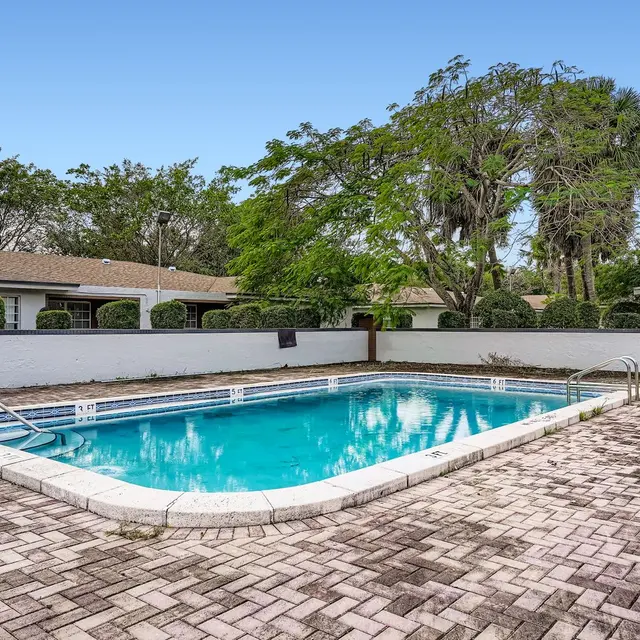 A residential swimming pool surrounded by a brick patio, set in a green landscape with trees in the background.