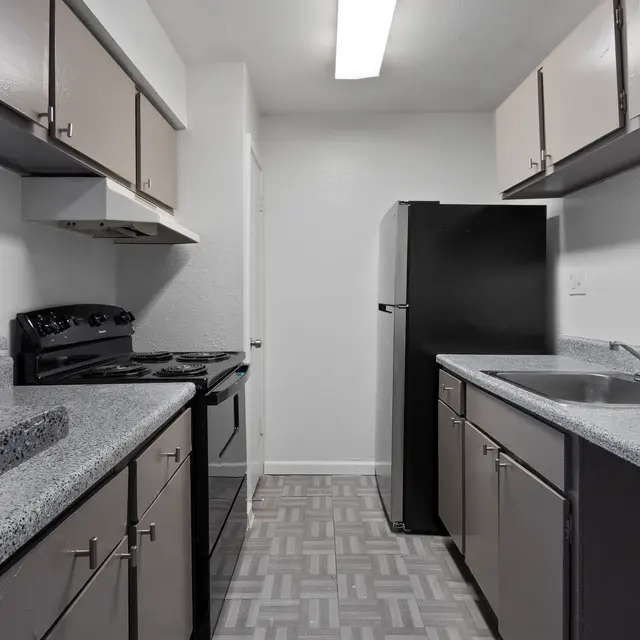 A modern kitchen featuring light-colored cabinets, a black refrigerator, and a black gas stove. The countertop is made of a speckled grey material. Floor tiles are arranged in a geometric pattern. A fire extinguisher is mounted on the wall.