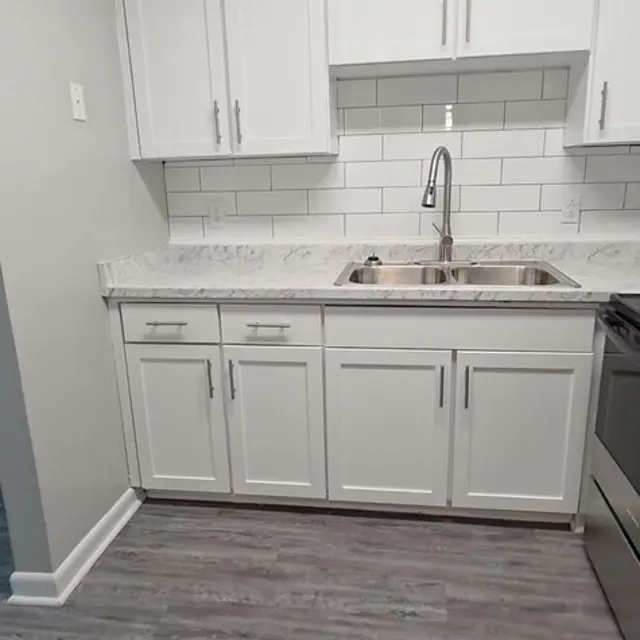 Modern kitchen with white cabinetry, a stainless steel sink, and marble countertops, featuring grey flooring and white tiled backsplash.