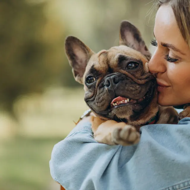 A woman gently kissing a dog while holding it in her arms outdoors. The dog, a French Bulldog, has a happy expression. They are surrounded by blurred greenery.