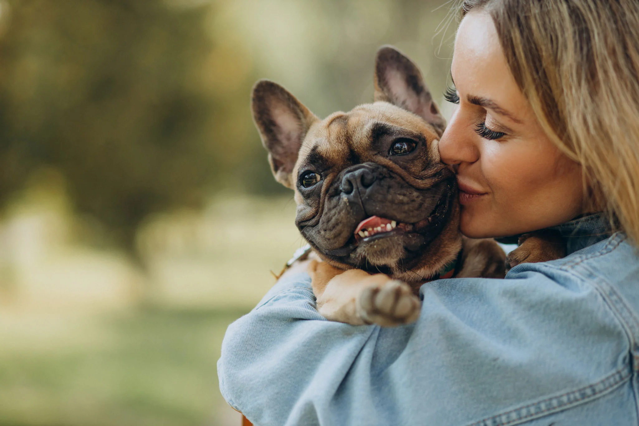 Joyful Connection: Woman and Her French Bulldog A woman gently kissing a dog while holding it in her arms outdoors. The dog, a French Bulldog, has a happy expression. They are surrounded by blurred greenery.