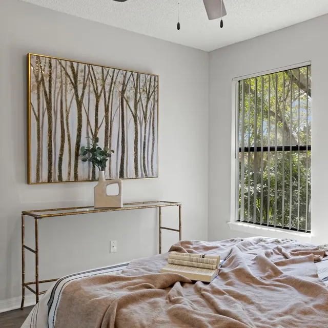 A well-decorated bedroom featuring a bed with a beige throw blanket, a modern ceiling fan, and a painting of trees on the wall. A small table with a plant and decorative object complements the natural light coming from the window.