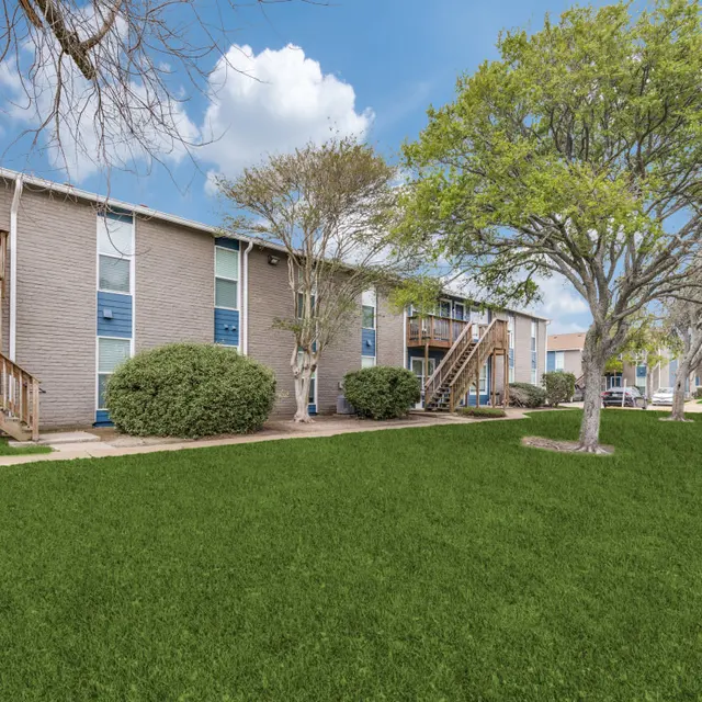 A view of an apartment complex with green lawns, trees, and a blue sky with clouds.