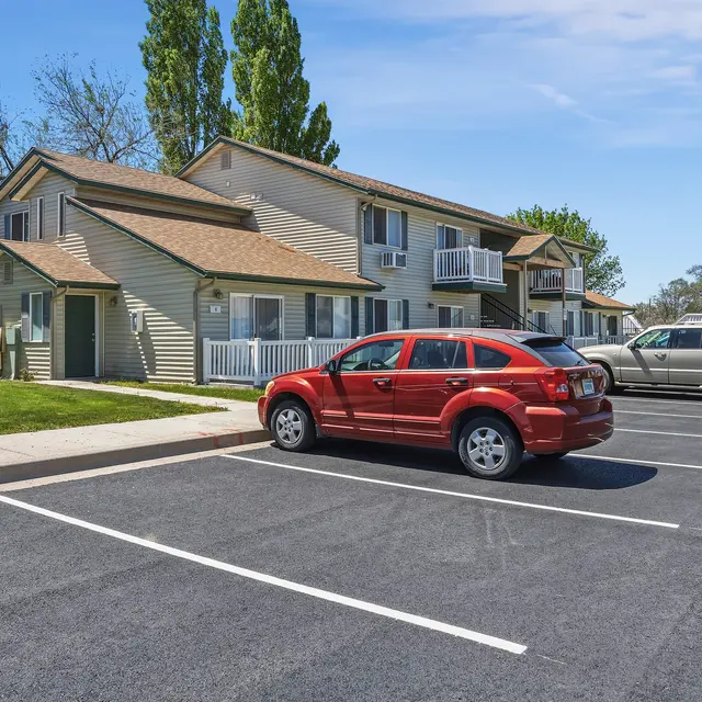 Apartment Complex with Parking View of a two-story apartment building with two parked cars, one red and one silver, in the foreground and green grass nearby.