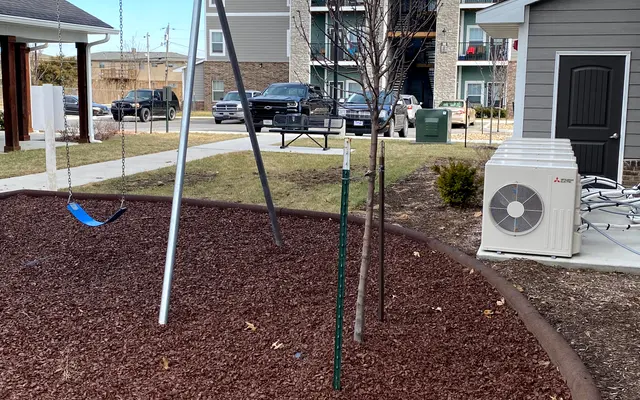 A playground area featuring a swing set on wood chip mulch, adjacent to an apartment complex. Trees are present in the background, and an air conditioning unit is visible nearby.
