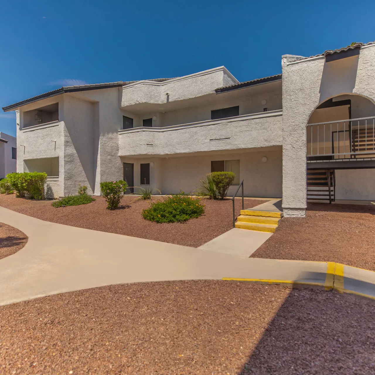 Exterior view of a multi-family apartment complex featuring two stories, a curved walkway, and landscaped areas with shrubs.